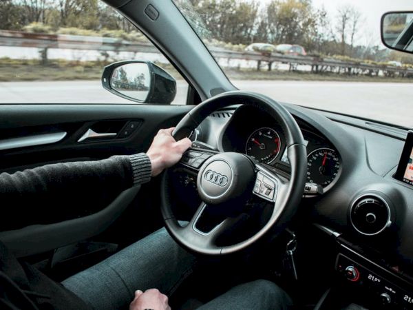A driver sits behind the wheel of a car (Audi), hands on the steering wheel, dashboard gauges and a navigation screen visible, driving on a highway.
