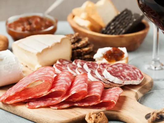 A charcuterie board featuring sliced cured meats, cheeses, bread, crackers, dips, nuts, and a glass of red wine.