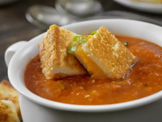 A warm bowl of tomato soup with crispy grilled cheese croutons on top, served in a white cup on a dark table.