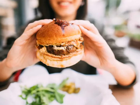 A person holds a juicy burger with greens on the side, ready to enjoy a tasty bite in a casual outdoor setting.