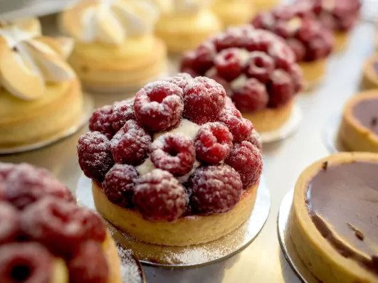A display of assorted pastries: raspberry-topped tarts, cream puffs, and shiny chocolate confections arranged in a bakery case.