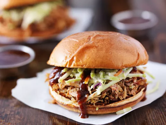 A pulled pork sandwich topped with coleslaw on a brioche bun, with a drizzle of barbecue sauce in the foreground and another sandwich in the background.
