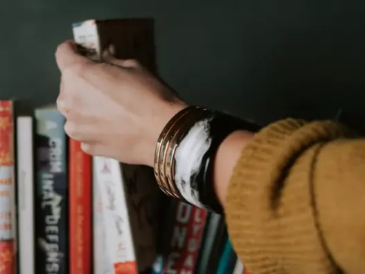 A person wearing a mustard sweater is browsing a bookshelf, gripping a book with several bracelets on their wrist.