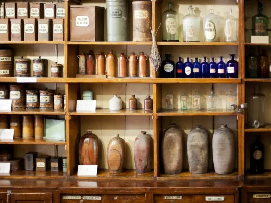A vintage apothecary shelf with numerous jars, bottles, and labeled containers arranged in wooden cubbies, evoking an old-time pharmacy atmosphere.