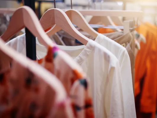 Clothes hanging on a rack in a boutique, pastel and bright tops on wooden hangers in a softly lit shop.