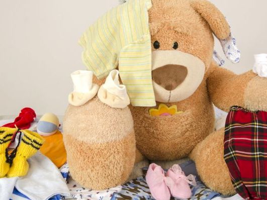 A pile of baby clothes and stuffed animals (teddy bears) with small shoes, hats, and a red plaid blanket, arranged on a bed.