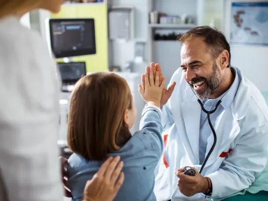A doctor high-fives a smiling child in a clinic, while a parent watches, fostering a warm, playful medical visit.