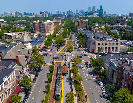 Aerial view of a city street lined with trees, buildings on both sides, a central tram line, and distant skyline on a clear day.