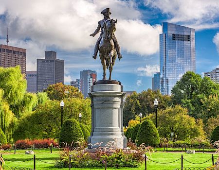 Statue of a rider on a tall pedestal in a park, with green trees, flowers, and a city skyline in the background under a blue, partly cloudy sky.