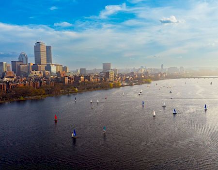 An aerial view of a city skyline along a river, with many sailboats on calm water and a bridge in the distance.