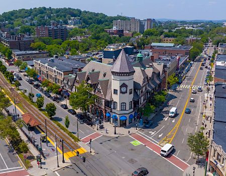 Aerial view of a small city center with a central clock tower building, streets, cars, and trees surrounding a busy intersection.