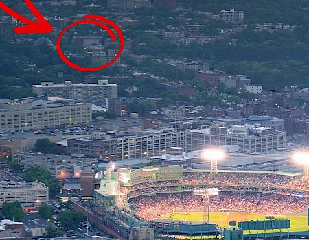 A cityscape with a stadium glowing at dusk; a distant circled area shows a faint tower or building behind trees, barely visible in the distance.