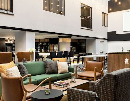 Hotel lobby lounge area with colorful sofas, modern chairs, coffee tables, and a reception desk; Arcadian Hotel signage visible. (approx 134 chars)