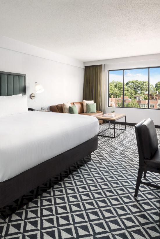 A modern hotel room with a large bed, bedside lights, a desk with TV, and a window overlooking trees, inviting a restful stay.