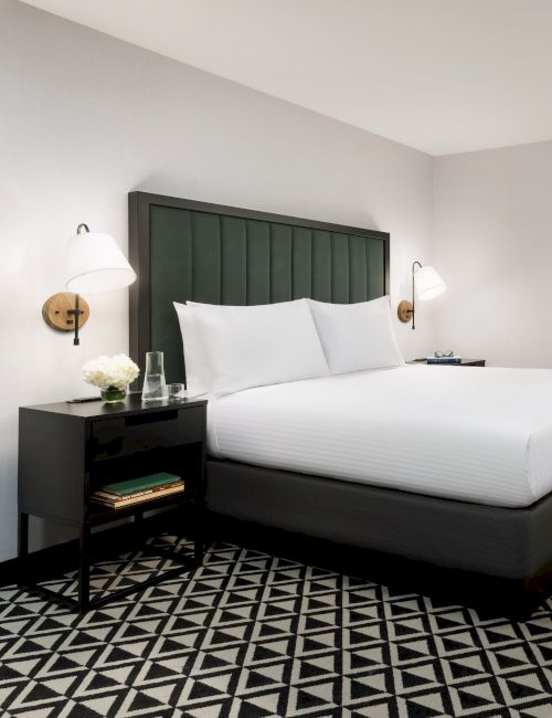 A modern hotel room: made bed with pillows, nightstand lamps, desk with chair and laptop, plus a wall TV.