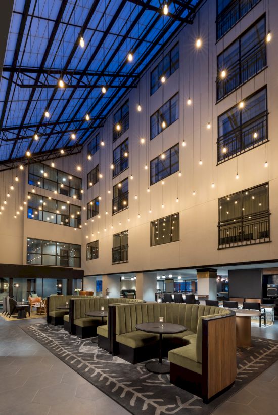 An elegant indoor atrium lounge with glass windows, warm pendant lights, and neatly arranged seating and tables, under a soaring steel-and-glass ceiling.