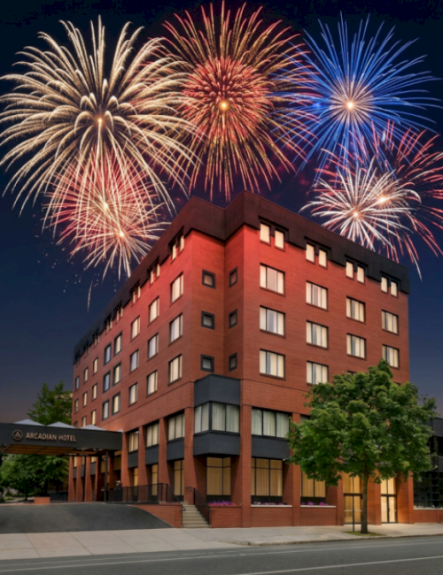 A red-brick hotel building is lit under colorful fireworks in the night sky.