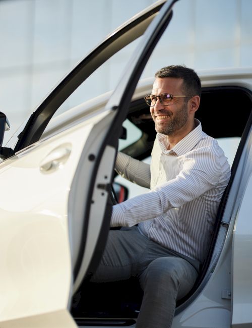 A smiling man in a striped shirt wearing glasses sits inside a car with the door open in bright daylight.