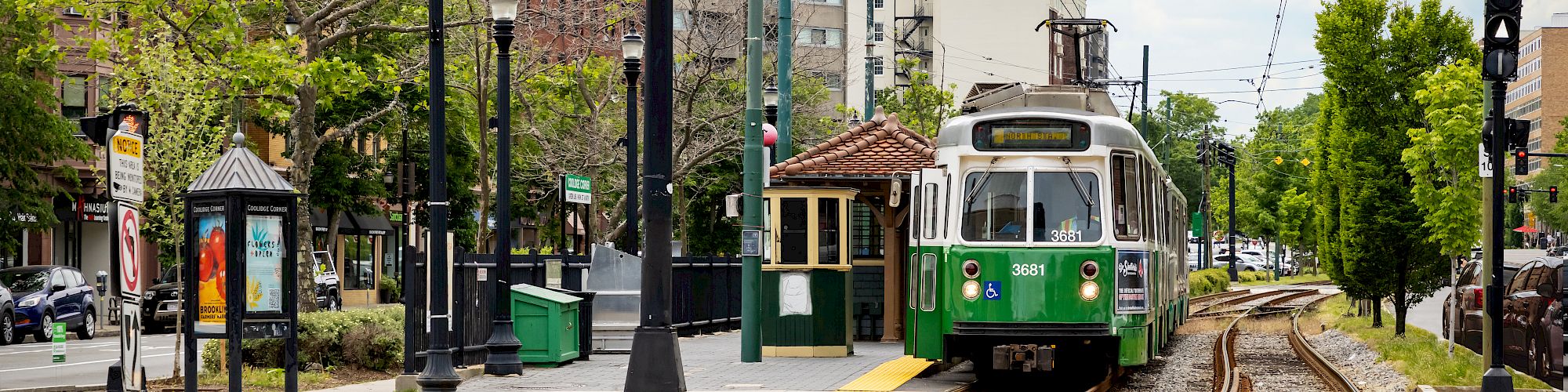 A green streetcar approaches along rail tracks in a city street, with American flags on poles and apartment buildings in the background.