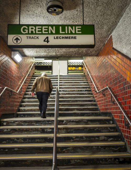A person climbs subway stairs toward a sign reading &ldquo;GREEN LINE, TRACK 4, LECHMERE&rdquo; in a brick tunnel.