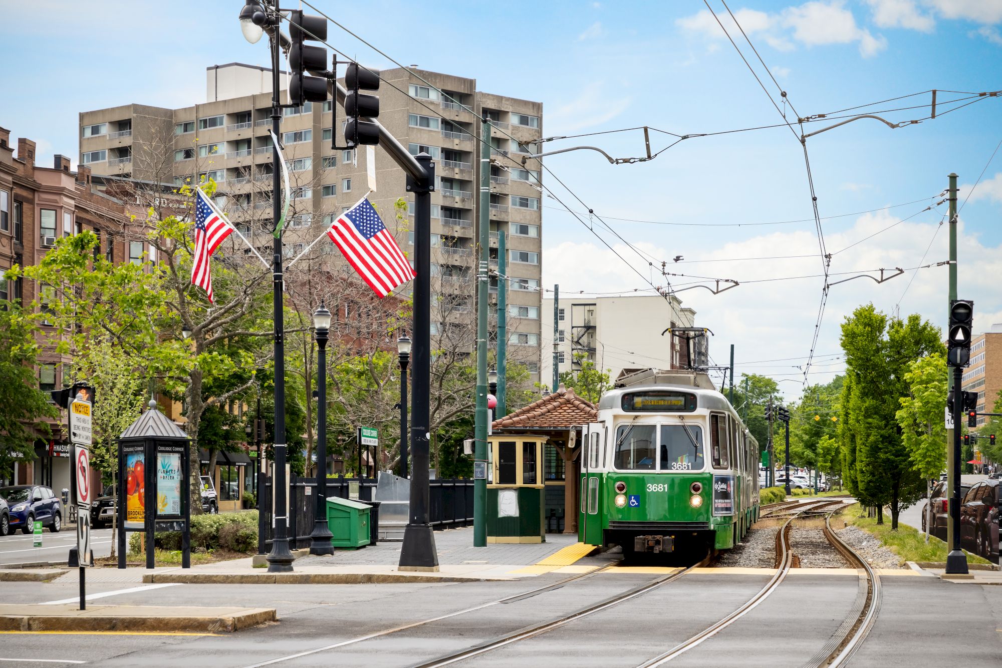A green-and-white streetcar waits on tracks under traffic signals, with American flags and apartment buildings in view.