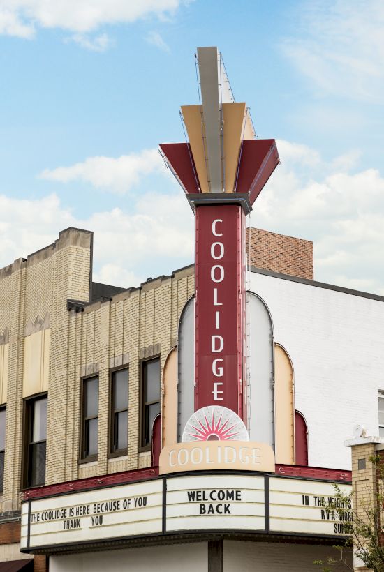 A theater marquee tower with &ldquo;COOLIDGE&rdquo; letters and an arched entrance sign on a city building under a blue sky.