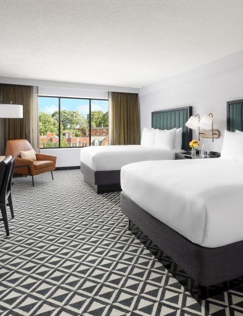 Modern hotel room with two beds, large windows, lamps, a desk, and a TV.