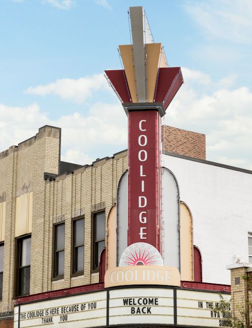 A street view of the Coolidge building facade, with a tall vertical &ldquo;COOLIDGE&rdquo; sign under a cloudy sky.