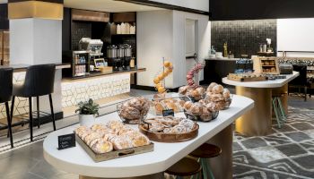 A hotel breakfast buffet with pastries and Danish/croissants, plus a coffee station and seating in a modern lounge.