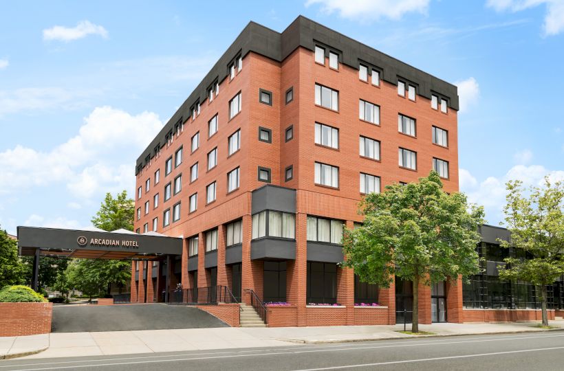 A red-brick multi-story hotel building with gray trim and the &ldquo;Arcadian Hotel&rdquo; sign in front under a blue sky.