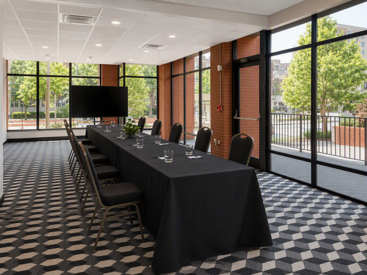 A conference room with a long table, black chairs, water glasses, brick walls, and large windows looking out on trees.