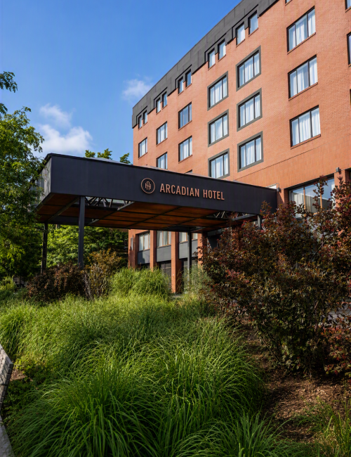 A modern Arcadian Hotel building with a covered entrance and sign, surrounded by greenery under a blue sky.