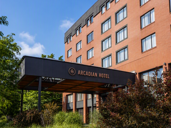 The Arcadian Hotel exterior shows a brick building, multi-story windows, and a dark canopy entrance under a blue sky.