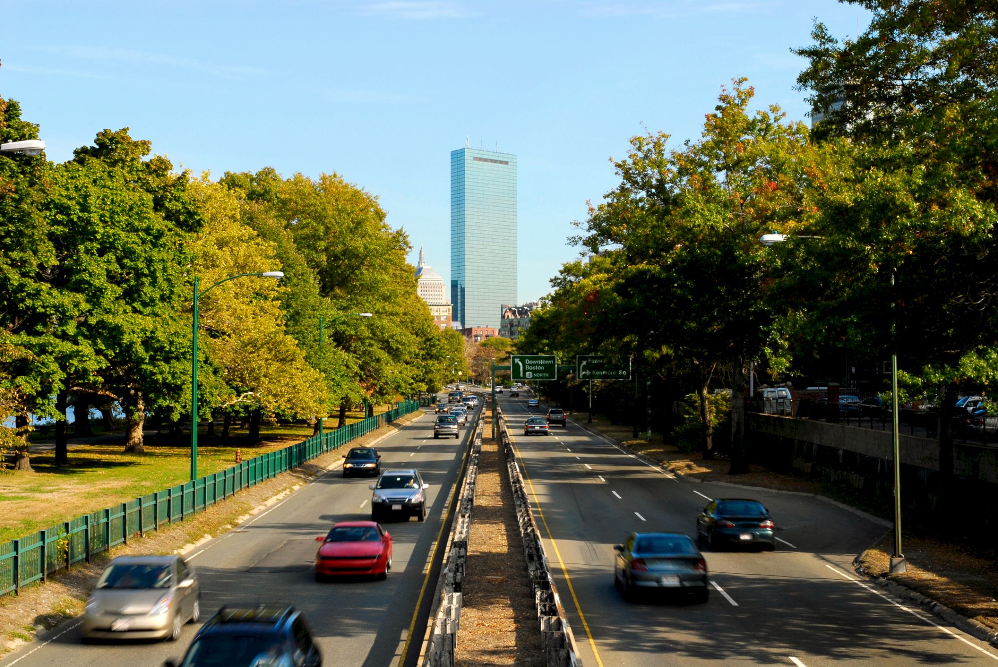 A busy tree-lined highway with cars heading toward a tall blue glass office tower in the downtown area.