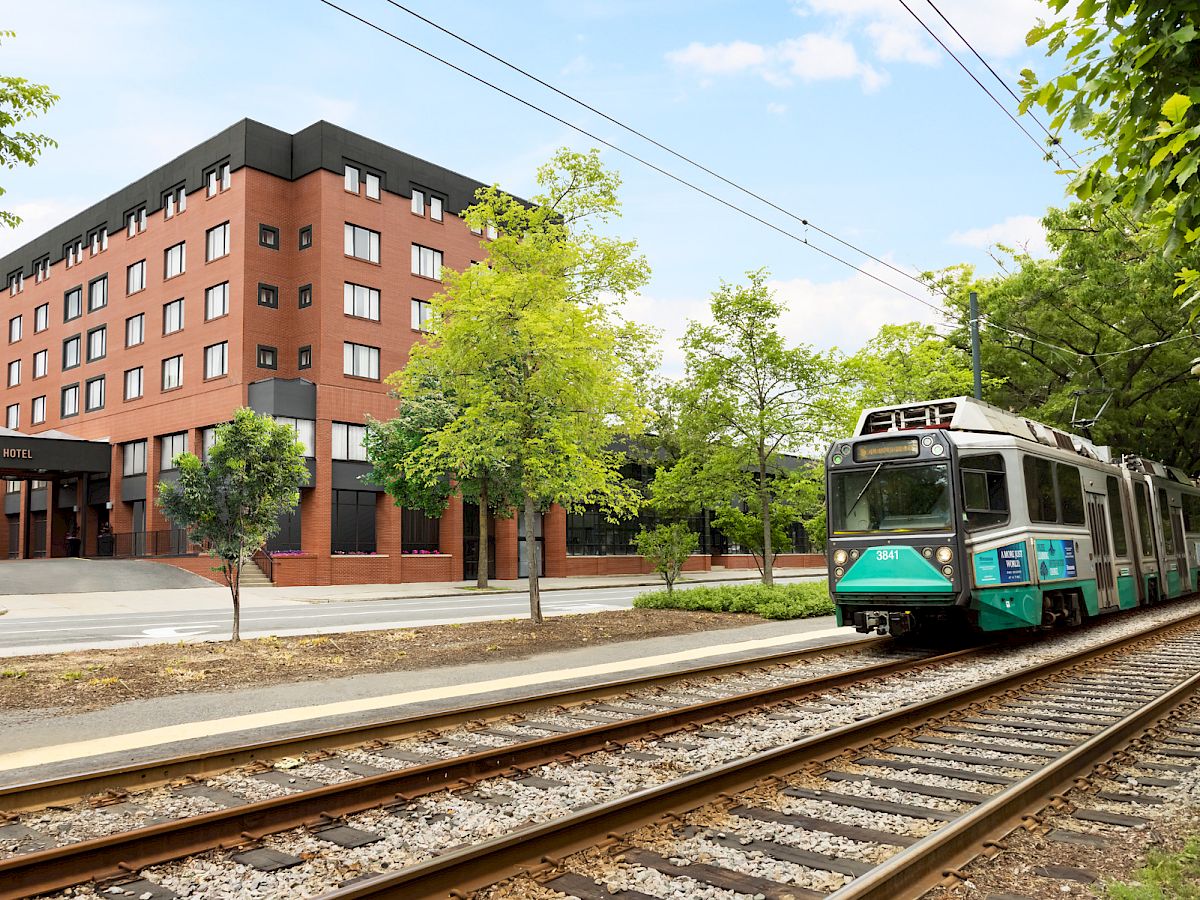 A green-and-white train rolls past the Arcadian Hotel as trees sway in daylight by the tracks.