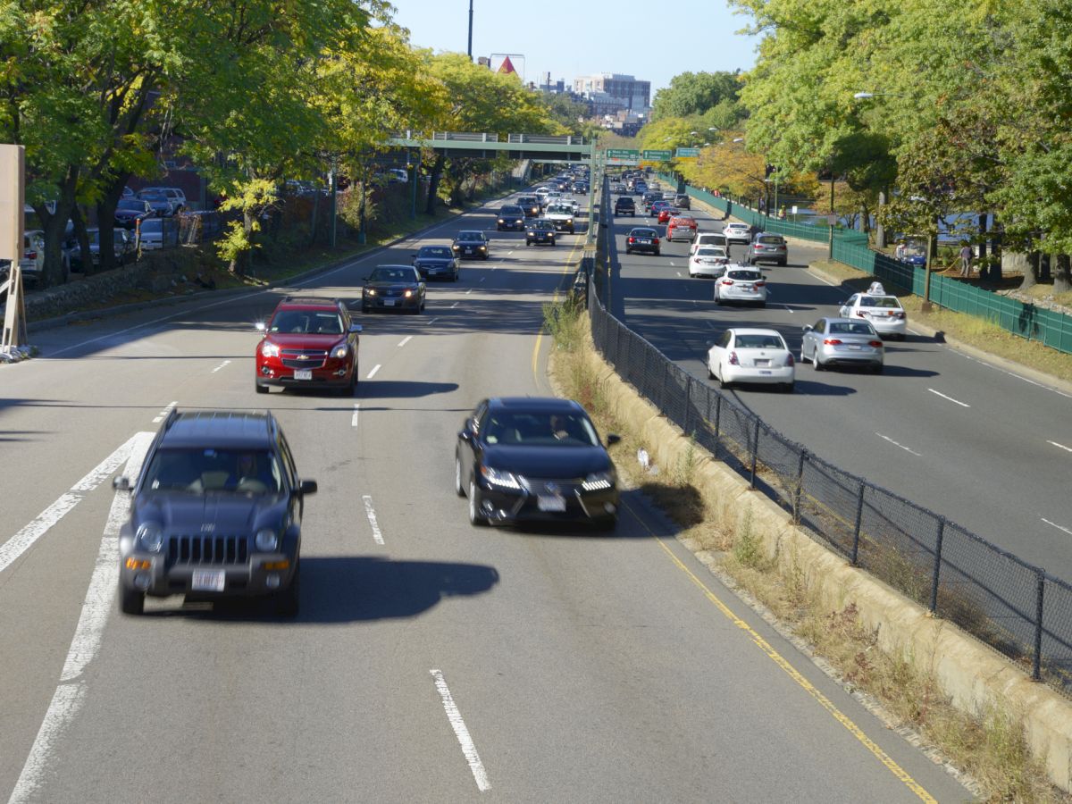 A busy multi-lane highway packed with cars, surrounded by green trees, with a city skyline visible in the distance.