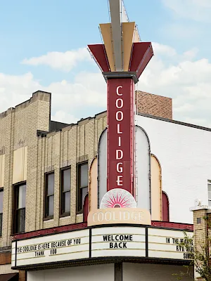 A vertical “Coolidge” theater sign rises above a marquee reading “Welcome Back” and “Coolidge,” under a blue sky.