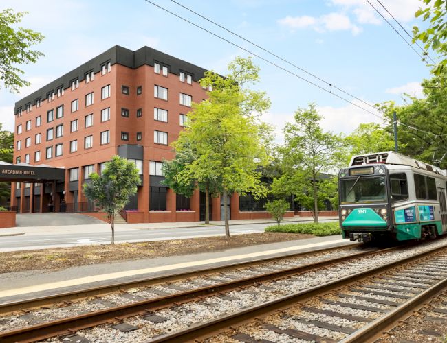 A red-brick Arcadian Hotel beside railway tracks as a green-and-white tram approaches through leafy trees.