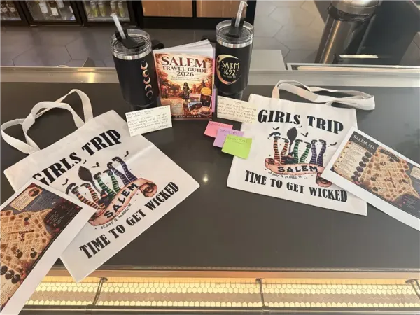 Salem, MA trip swag: two tote bags reading “Girls Trip—Time to Get Wicked,” travel guides, stickers, and drink cups on a table.