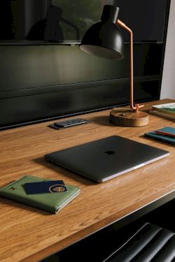 A desk setup: a black lamp with a copper stand, a laptop, a phone, a notebook, and books by a window.