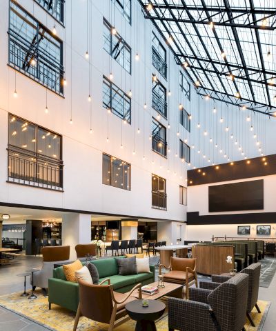 A modern hotel atrium with cozy lounge seating, warm string lights, and large windows in a glass-roof courtyard ending.