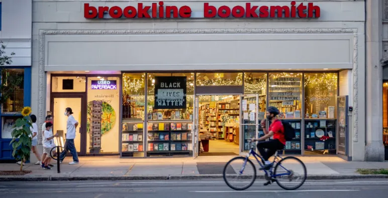A storefront of brookline booksmith with glowing lights, books, pedestrians, and a cyclist outside.