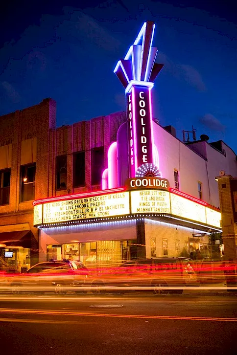 A neon-lit theater marquee labeled “Coolidge” at night, with city light trails and a tall vertical sign.