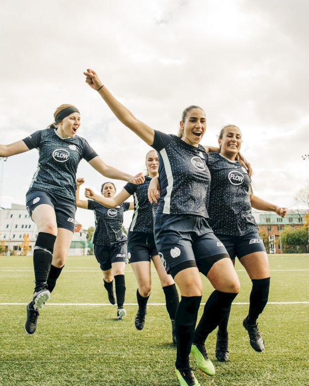 A women&rsquo;s soccer team celebrates on the field, shouting and jumping in matching kits under a cloudy sky.
