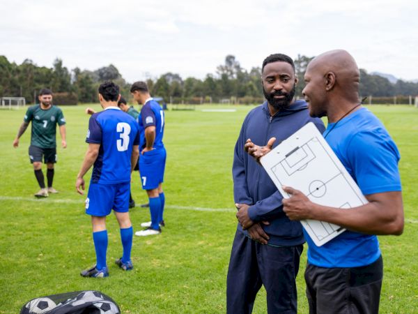 A soccer coach holds a tactics board while players in blue and green practice drills on the field.