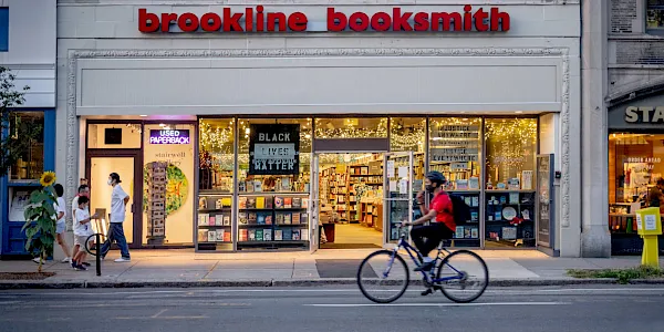 A storefront for Brookline Booksmith bookstore, with lit windows, posters, pedestrians, and a bicycle outside.