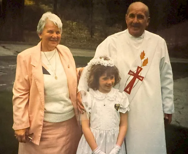 An elderly couple poses with a young child on their lap, all in white outfits featuring a red cross and cheerful smiles.